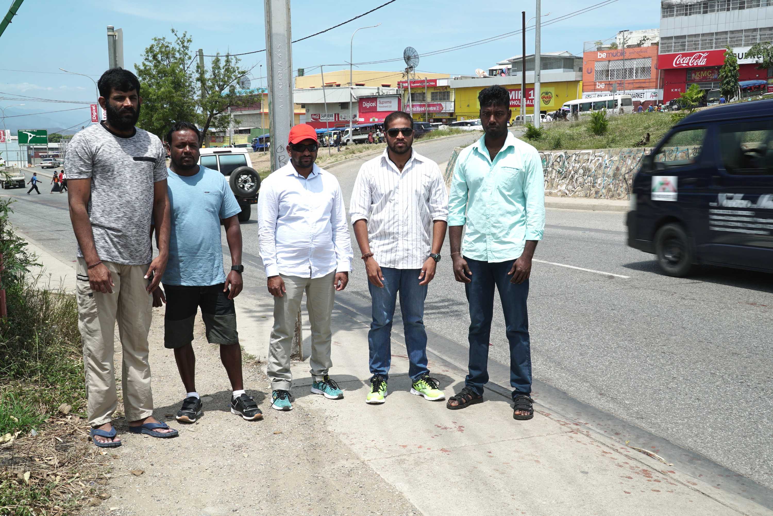 Sri Lankan refugees stand on a sidewalk by a busy road in Port Moresby, there are shops behind them.