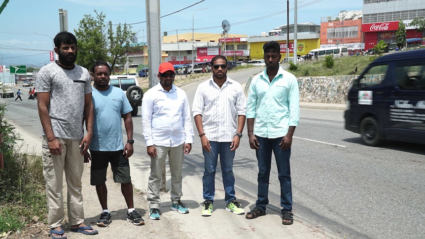 Sri Lankan refugees stand on a sidewalk by a busy road in Port Moresby, there are shops behind them.