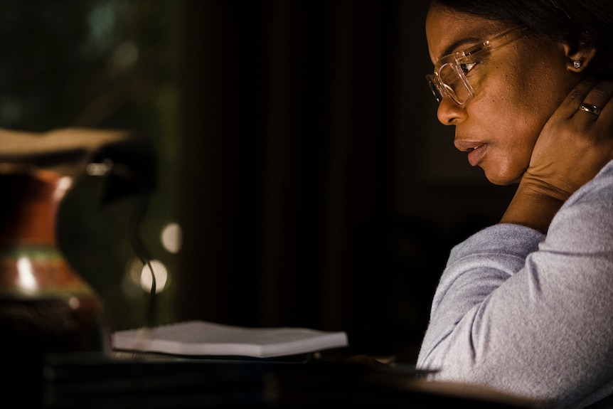 A middle-aged Black woman wearing a pale blue knit and clear glasses sits at a desk reading papers by lamplight.