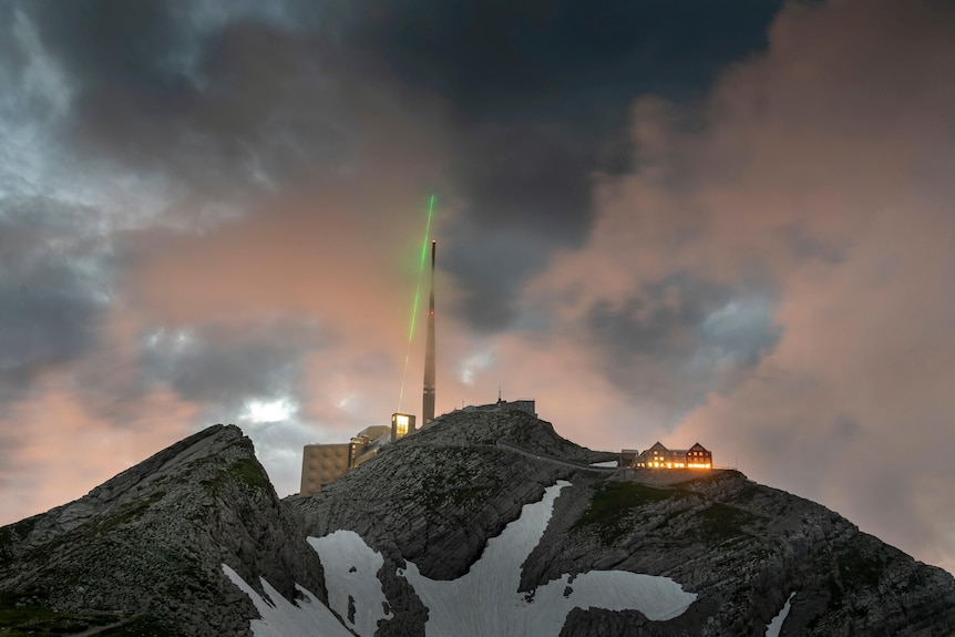 A mountain with snow on it is pictured, a green laser beam comes out of a high structure. 