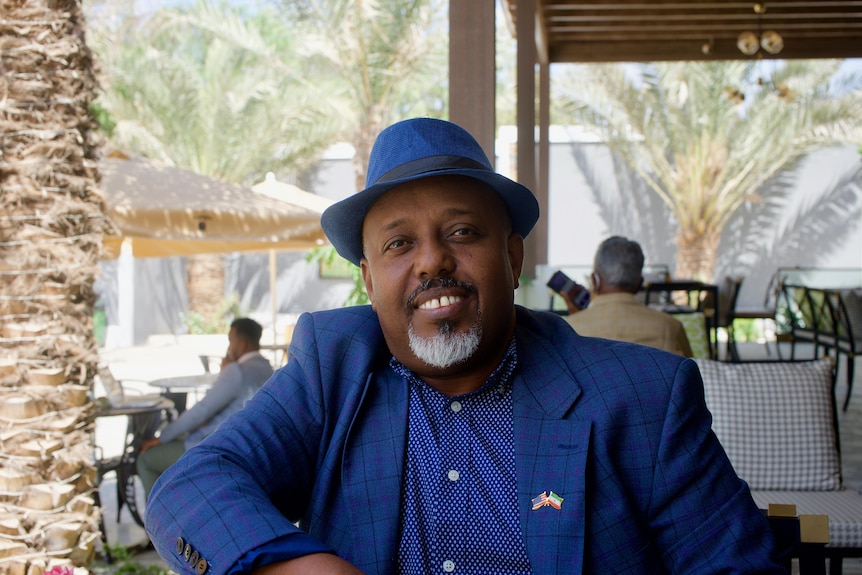 Man in a blue shirt, jacket and hat with his arm resting on a chair smiles widely.