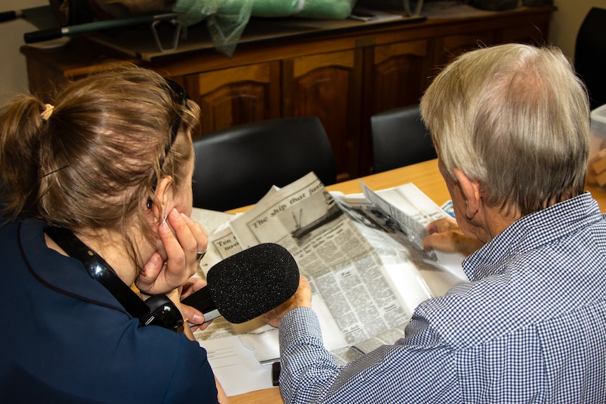 A women with a microphone interviews and older man as he pores over old newspaper clippigs.
