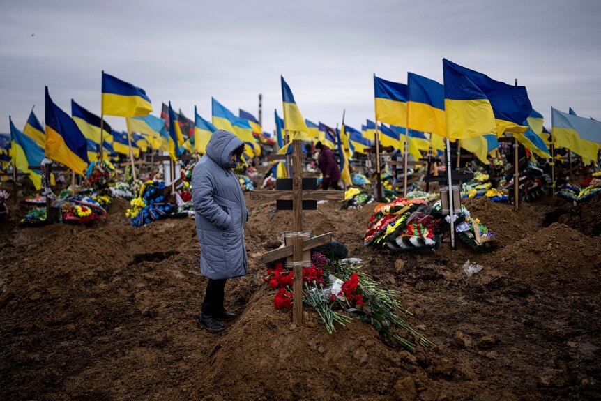 A wife looks at the grave of her husband as Ukraine flags a seen flying over other graves behind her.