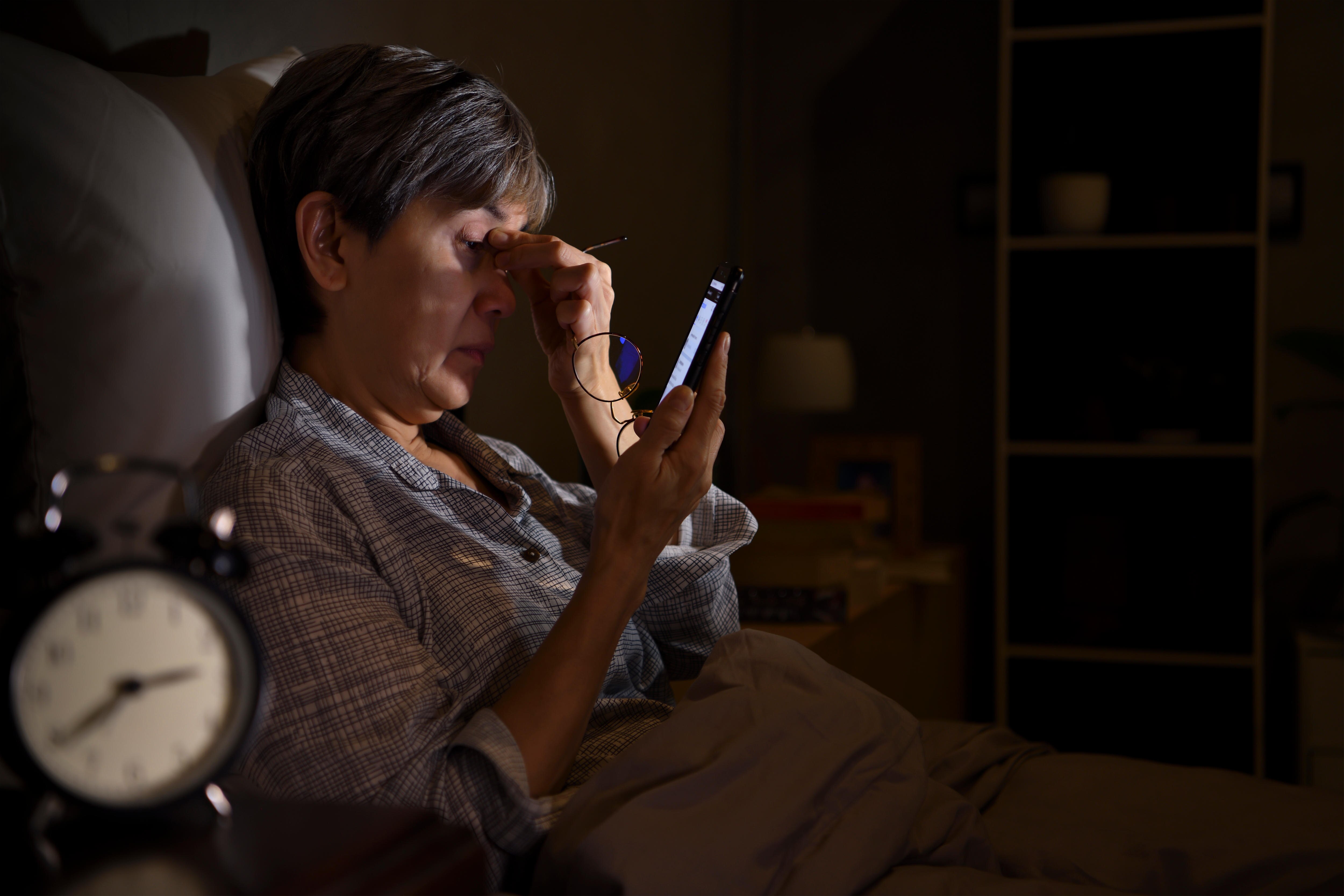 And older woman with sore and tired eyes, using a smartphone while lying in bed late at night.