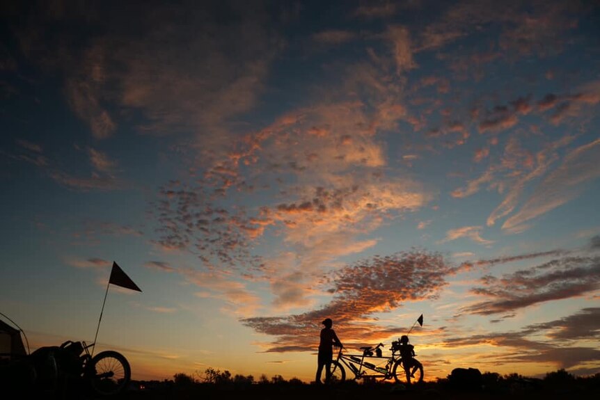 Orange clouds at sunset with silhouettes of two people and two bicycles in the foreground.