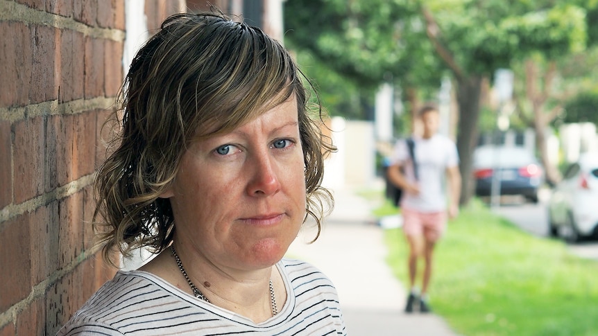 A woman looks at the camera and behind her is a street with a blurry man