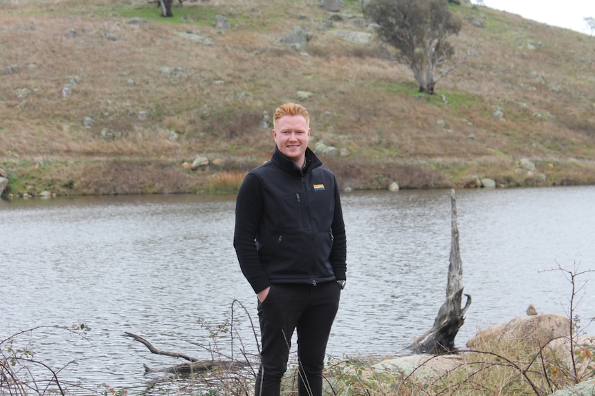 A man wearing a black vest and black trousers stands in front of a dam.