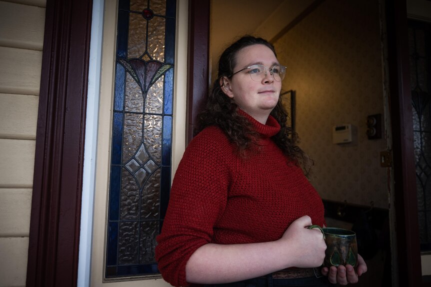 A white woman with curly brown hair and glasses, stands at her front door
