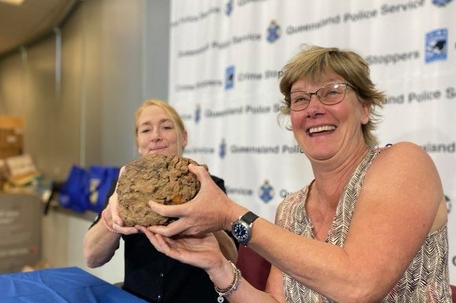Two women hold up a soccer-ball sized meteorite sitting in front of a police banner