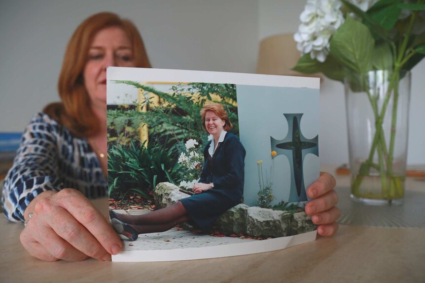 Woman holds an old photograph of a young girl with red hair sitting in uniform next to a cross.