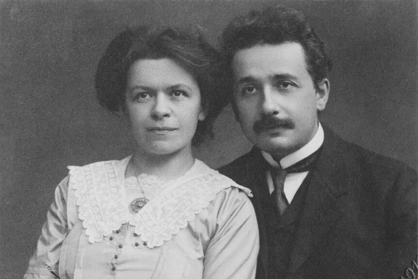 Black and white photo of young woman in white dress and man with moustache sitting side by side.