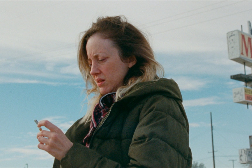 A film still of Andrea looking at her cigarette as she stands on the street.