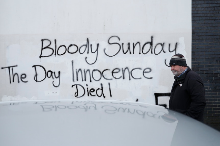 Un bărbat trece pe lângă un perete cu un slogan pictat cu spray „Bloody Sunday The Day Innocence Died” 