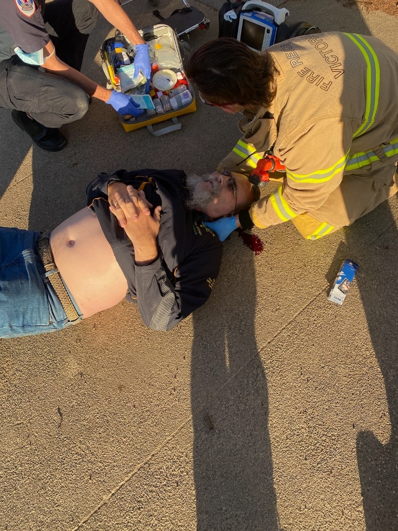 Man laying on the ground injured with an emergency responder cradling his head. There is a small pool of blood next to his head.