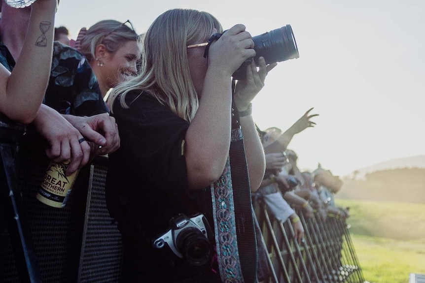 Ruby stands against a steel crowd barrier and holds a large professional camera up to her face.