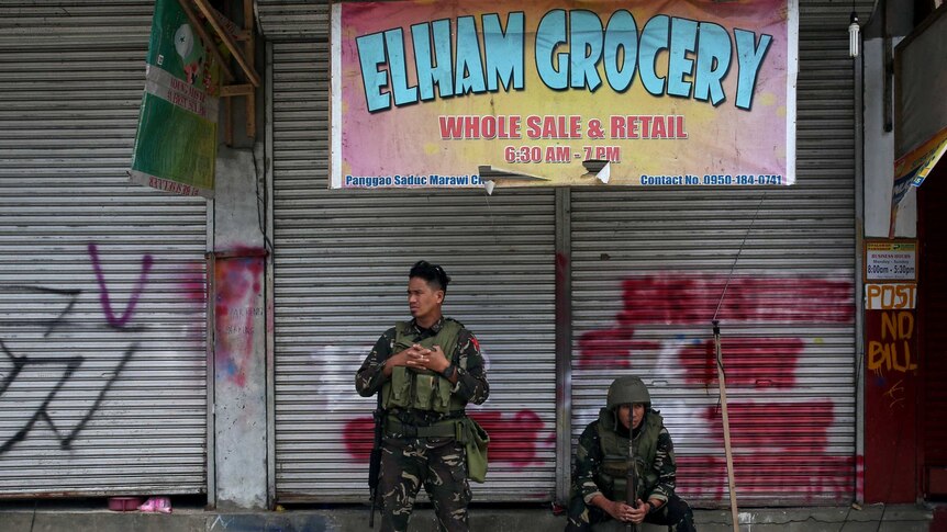 Two soldiers, one sitting and another standing, rest in front of a closed grocery store.