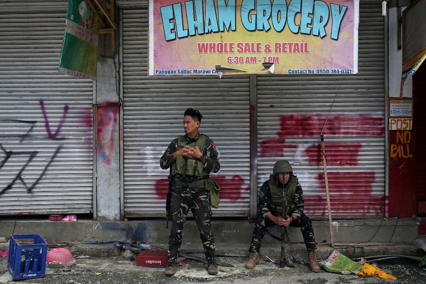 Two soldiers, one sitting and another standing, rest in front of a closed grocery store.