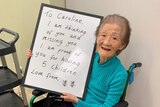 Older woman sitting in wheelchair holds up sign addressed to her daughter, saying she is missing her