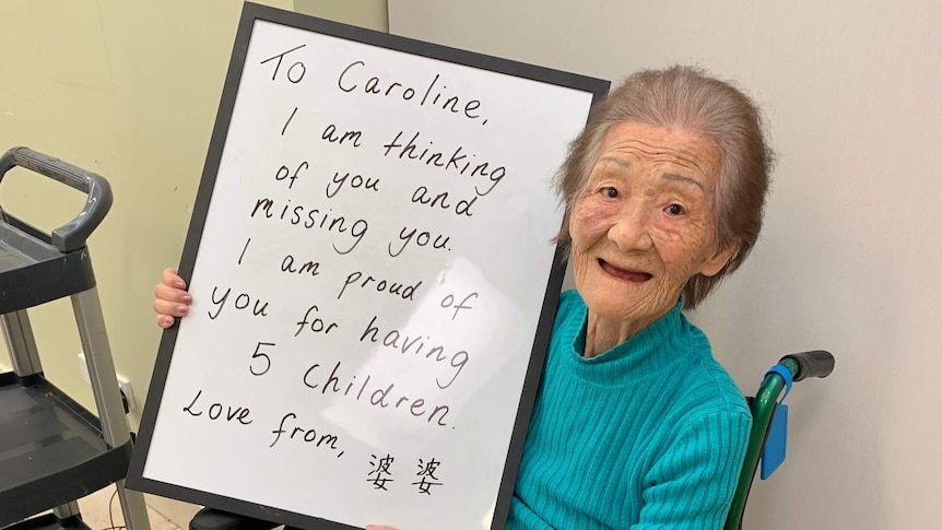 Older woman sitting in wheelchair holds up sign addressed to her daughter, saying she is missing her