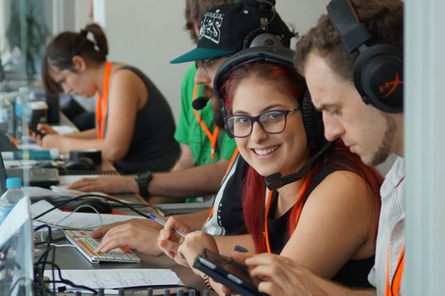 A young woma, seated at a desk among other broadcast producers smiles at the camera.