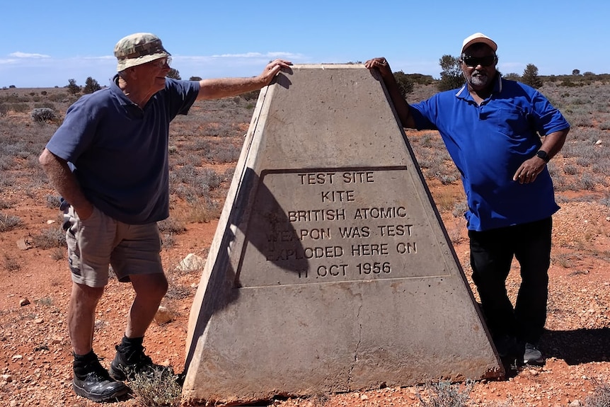 Two men stand either side of triangular prism shaped memorial that reads "british atomic weapon was test exploded here in octob