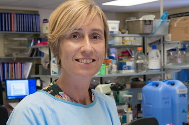 Professor Claudine Bonder, in blue lab coat and rubber gloves, holding a test tube in a laboratory