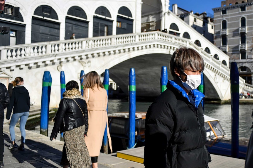 A man with dark hair and winter clothes walks along a promenade on a sunny day in front of a bridge.