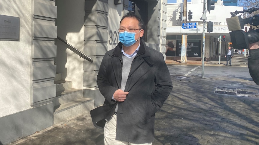 A man with black hair and glasses wearing a face mask and coat on a city street