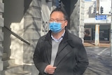 A man with black hair and glasses wearing a face mask and coat on a city street