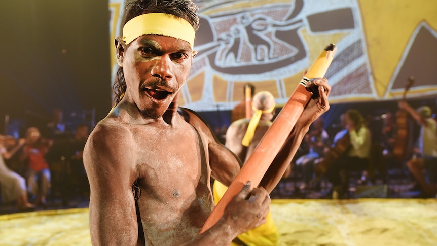 Young man with ochre on face and torso, wearing yellow head-band, mugs for camera while playing wooden instrument as if guitar.