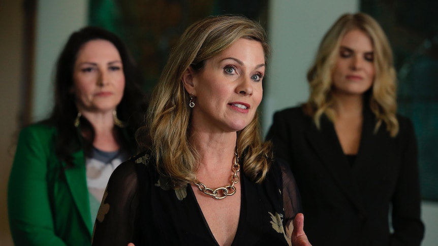 A blonde woman with shoulder length hair speaking with two other women behind her on either side