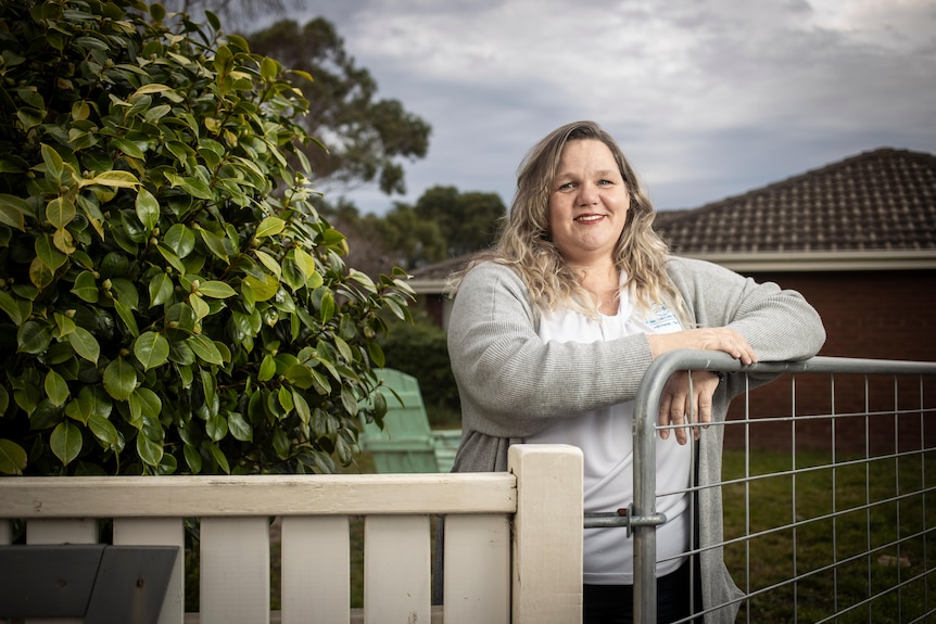 A woman wearing a pale shirt leans over the front of her fence line, smiling.