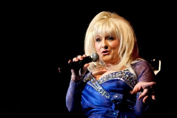 A woman with a blonde wig holding a microphone and dressed in a blue dress.