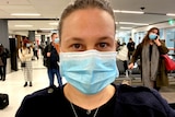 A close-up shot of a woman wearing a face mask with airline passengers standing in the background.