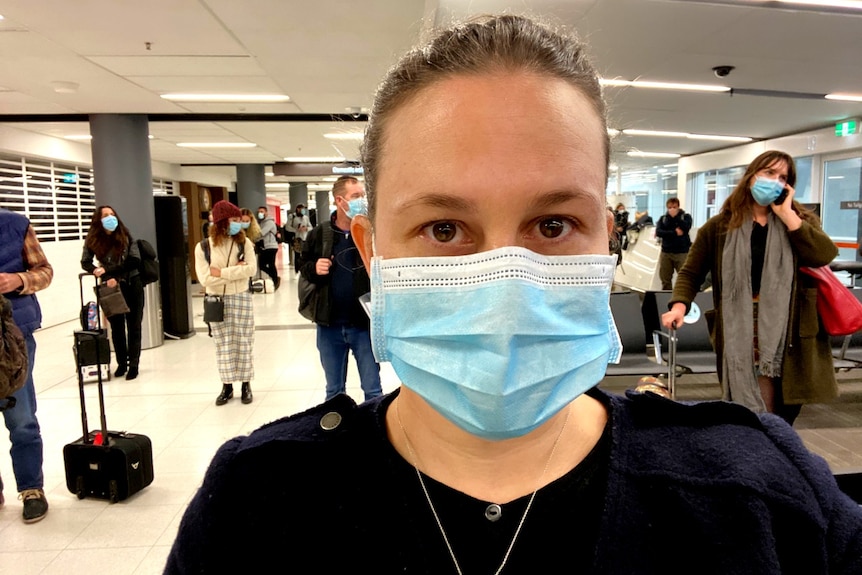 A close-up shot of a woman wearing a face mask with airline passengers standing in the background.