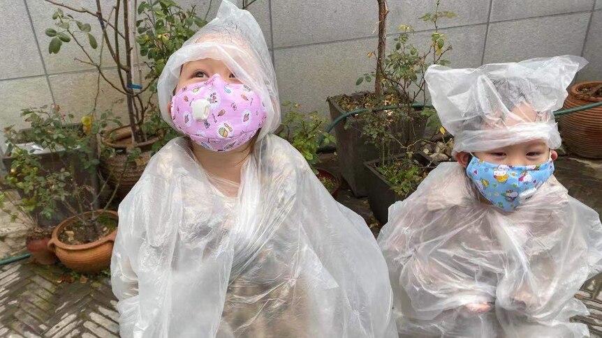 Two toddlers stand in a courtyard covered in plastic, wearing face masks.