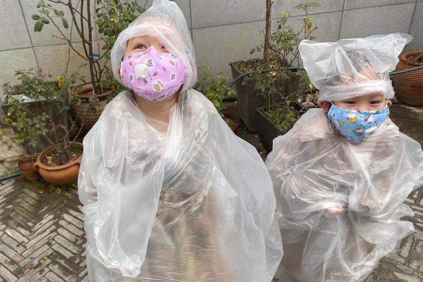 Two toddlers stand in a courtyard covered in plastic, wearing face masks.