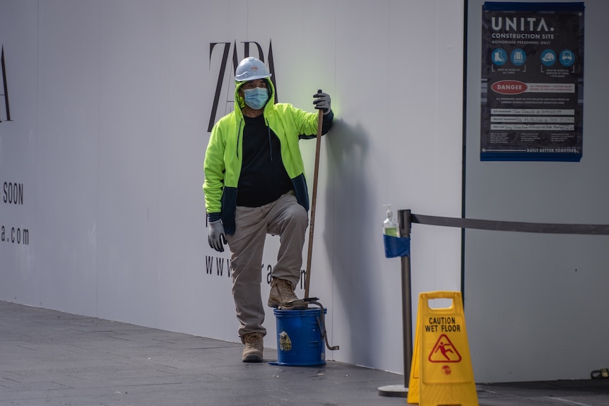 A man in a mask stands with his foot on a bucket and holding a mop