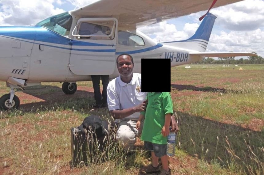 A dark-skinned man crouches near a blue and white aeroplane, posing with an unidentified child.