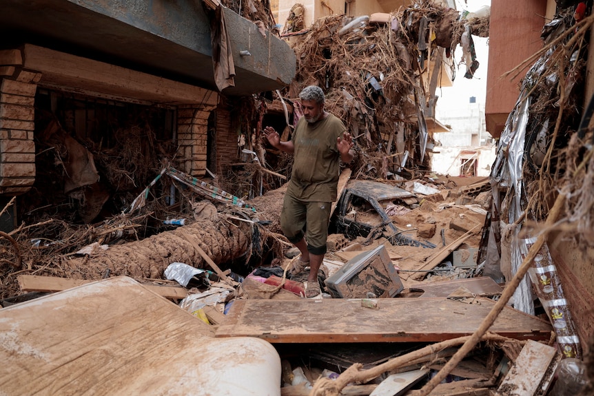 A man lifts his hands with palms facing forward as he steps over rubble in a street filled with debris