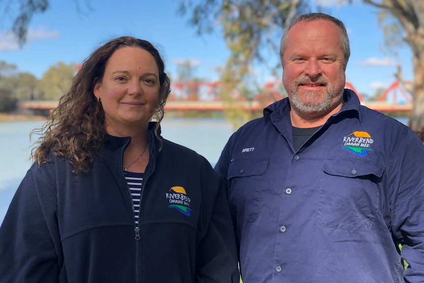 A woman and a man stand on the banks of the River Murray with a red bridge just visible in the distance