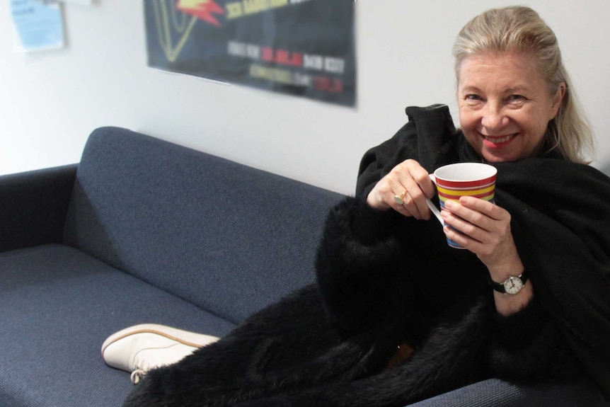 On a blue couch, a woman wrapped in black jacket and black scarf holds a mug hear her face and smiles widely.