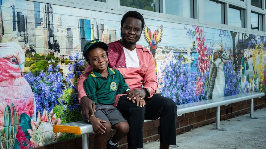 Victor sits on a bench with his young son King George, both smling in front of a colourful painted mural.