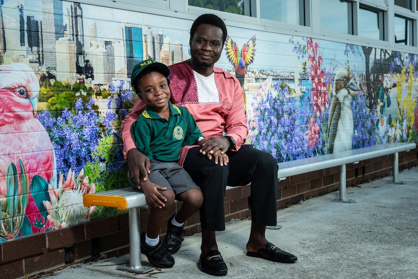 Victor sits on a bench with his young son King George, both smling in front of a colourful painted mural.