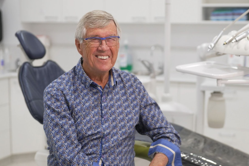 A dentist with grey hair and wearing glasses smiles widely sitting near his dentist's chair.