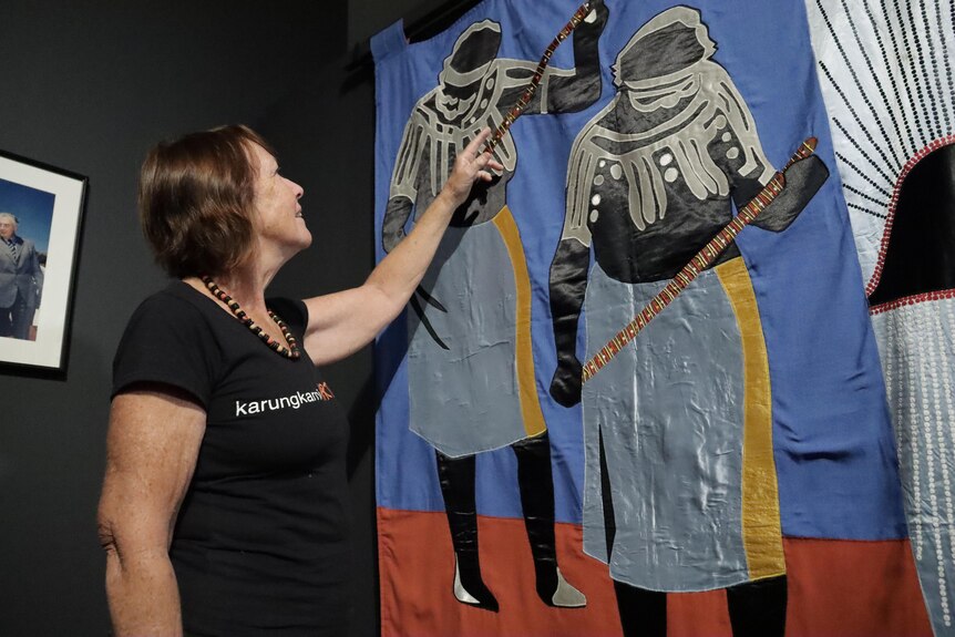 A woman in a black t-shirt points to a textile mural depicting Aboriginal women in traditional body paint