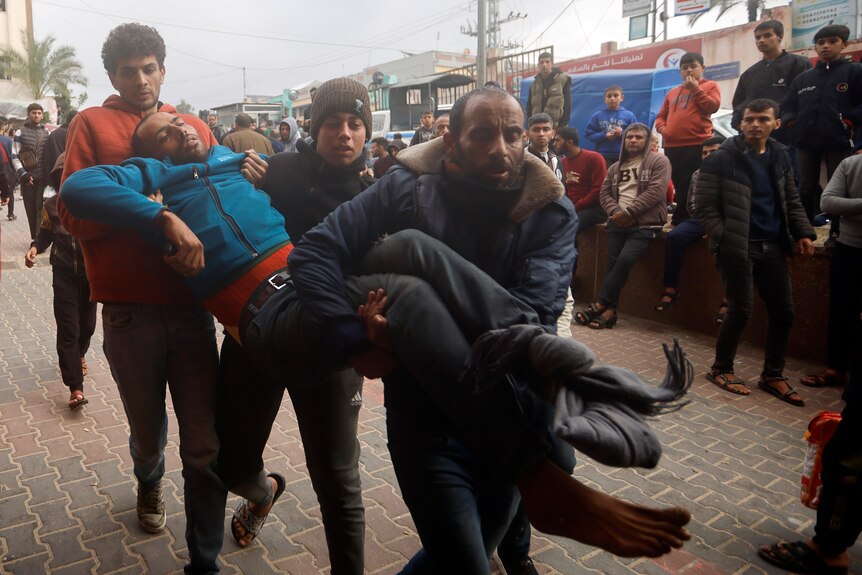 Three men carry a man in blue jumper into a hospital