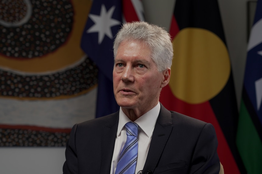 Smith sits in front of Australian, Aboriginal and Torres Strait flags, his hands raised in gesture as he speaks