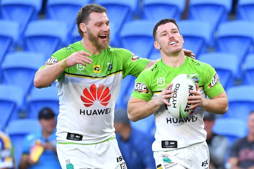 A Canberra Raiders NRL player holds the ball in two hands as a teammate smiles and approaches him to congratulate him.
