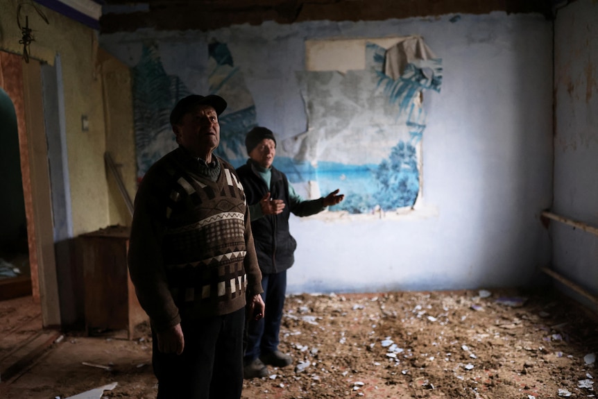 Two people stand inside a house and look up at the walls which are damaged. 